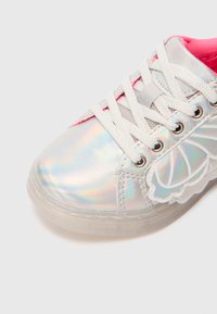 Primer plano de una zapatilla blanca iridiscente con cordones blancos y suela translúcida, que presenta un detalle en forma de ala y un forro interior rosa.