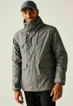 Mężczyzna uśmiechający się, ubrany w szarą zimową kurtkę z kapturem i dużymi kieszeniami oraz ciemną czapkę z daszkiem, stojący na jednolitym tle.