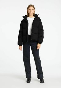 Sort puffjakke med hætte, med et quiltet design og trykknapper. Bæres over en hvid skjorte og parret med mørke jeans og støvler.