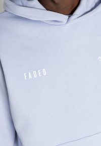 Felpa di colore azzurro chiaro con cappuccio regolabile, texture morbida e logo bianco riflettente "FADED" sul petto. Design moderno e informale.