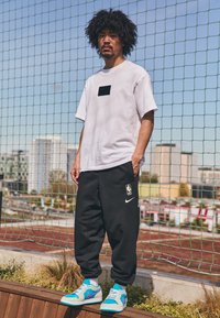 Witte t-shirt met korte mouwen en een zwarte patch, zwarte trainingsbroek met een wit Nike-logo en NBA-insigne, en kleurrijke sneakers.