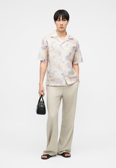 Homme portant une chemise à manches courtes à boutons à motif floral, un pantalon beige à jambes larges, des sandales noires, et tenant un sac à main noir sur un fond blanc.