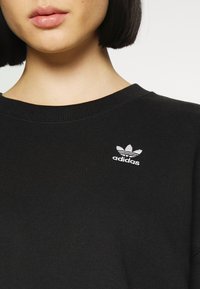 Melns krekls ar kapuci, izgatavots no kokvilnas, ar ribotu apkakli un mazu balto Adidas logo izšūtu uz krūšu daļas.