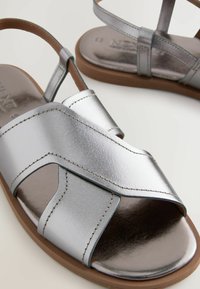 Sandali metallici argento con design a fascia incrociata, punta aperta e texture liscia, con suola marrone e dettagli di cucitura.