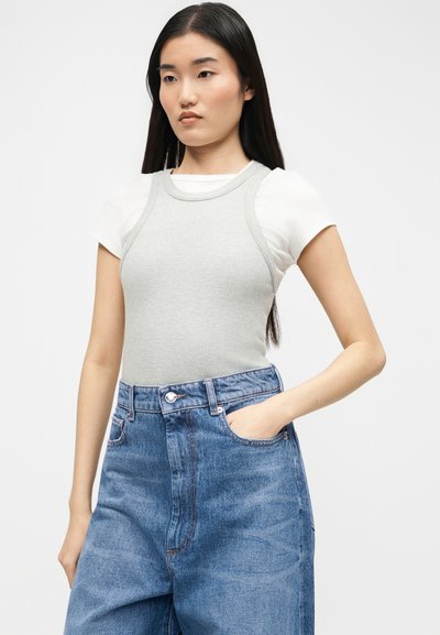 Νεαρή γυναίκα με μακριά σκουρόχρωμα μαλλιά, που φοράει λευκό κοντομάνικο πουκάμισο κάτω από ένα ανοιχτό γκρι αμάνικο τοπ και ψηλόμεσα μπλε τζιν.