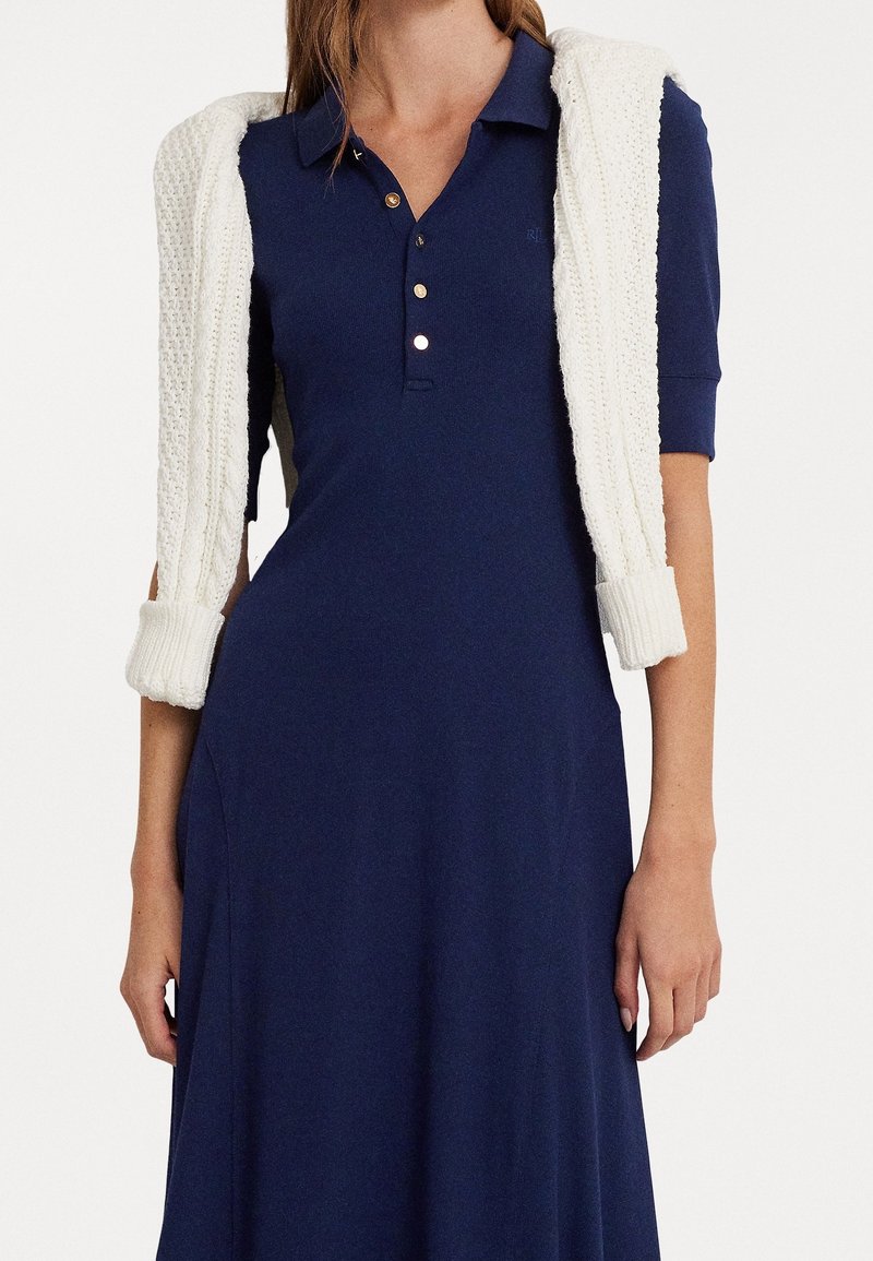 Mörkblå polo klänning med en figurnära silhuett, korta ärmar och tre gyllene knappar. En vit stickad tröja är draperad över axlarna.