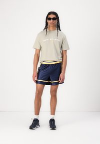 T-shirt vert clair avec logo "New Balance", shorts bleu marine avec accents jaunes et baskets sombres. Le modèle porte des lunettes de soleil noires.