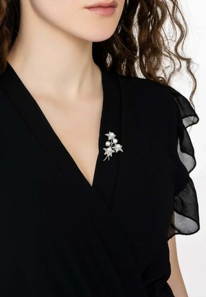 Black wrap dress with sheer fabric sleeves, featuring a silver floral brooch with pearls and clear accents pinned to the chest area.