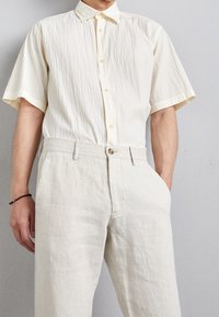 Beżowa koszula z krótkim rękawem o teksturowanej powierzchni, zapinana na guziki z przodu, w duecie z jasnymi lnianymi spodniami. Dominują subtelne, naturalne odcienie.