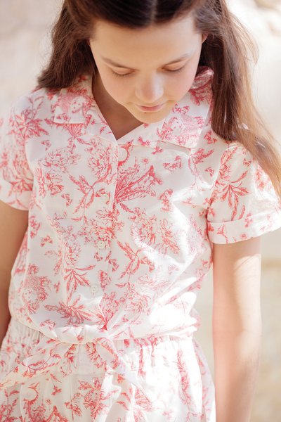 Weißes Kleid mit roten Blumenmustern, kurzen Ärmeln, Kragen und Bindegürtel in der Taille. Hergestellt aus leichtem Stoff mit glatter Textur.