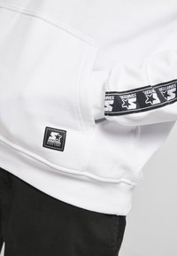 Felpa bianca con patch del logo Starter nero sulla tasca e striscia del logo nera sulla manica, abbinata a pantaloni neri.