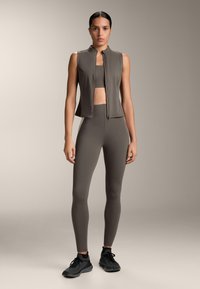 Vrouw staand, gekleed in een taupekleurig mouwloos vest met ritssluiting, een bijpassende sportbeha en high-waisted leggings, met zwarte sportschoenen, op een neutrale achtergrond.