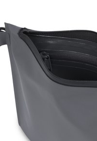 Bolso gris con cremallera y textura suave y mate. Cuenta con bolsillos interiores y una forma redondeada con bordes cosidos.