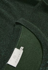 Tissu tricoté vert foncé pailleté avec une étiquette blanche indiquant le nom de la marque et la taille M près d'un bord d'encolure incurvé.