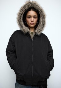 Czarna pikowana kurtka z kapturem wyściełanym sztucznym futrem, z przednim zamkiem błyskawicznym, bocznymi kieszeniami i ściągaczami na mankietach. Tekstura wygląda na gładką i miękką.