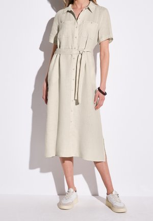 Femme portant une robe midi beige à boutons, manches courtes, avec une ceinture à la taille et des baskets blanches, debout devant un fond uni clair.