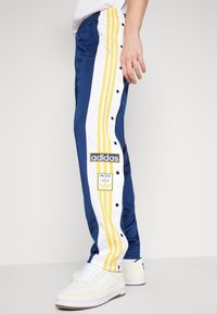 Blå träningbyxor med vita och gula sidestreck, tryckknappar och Adidas-logotyp. Tyget är glatt och något stretchy.