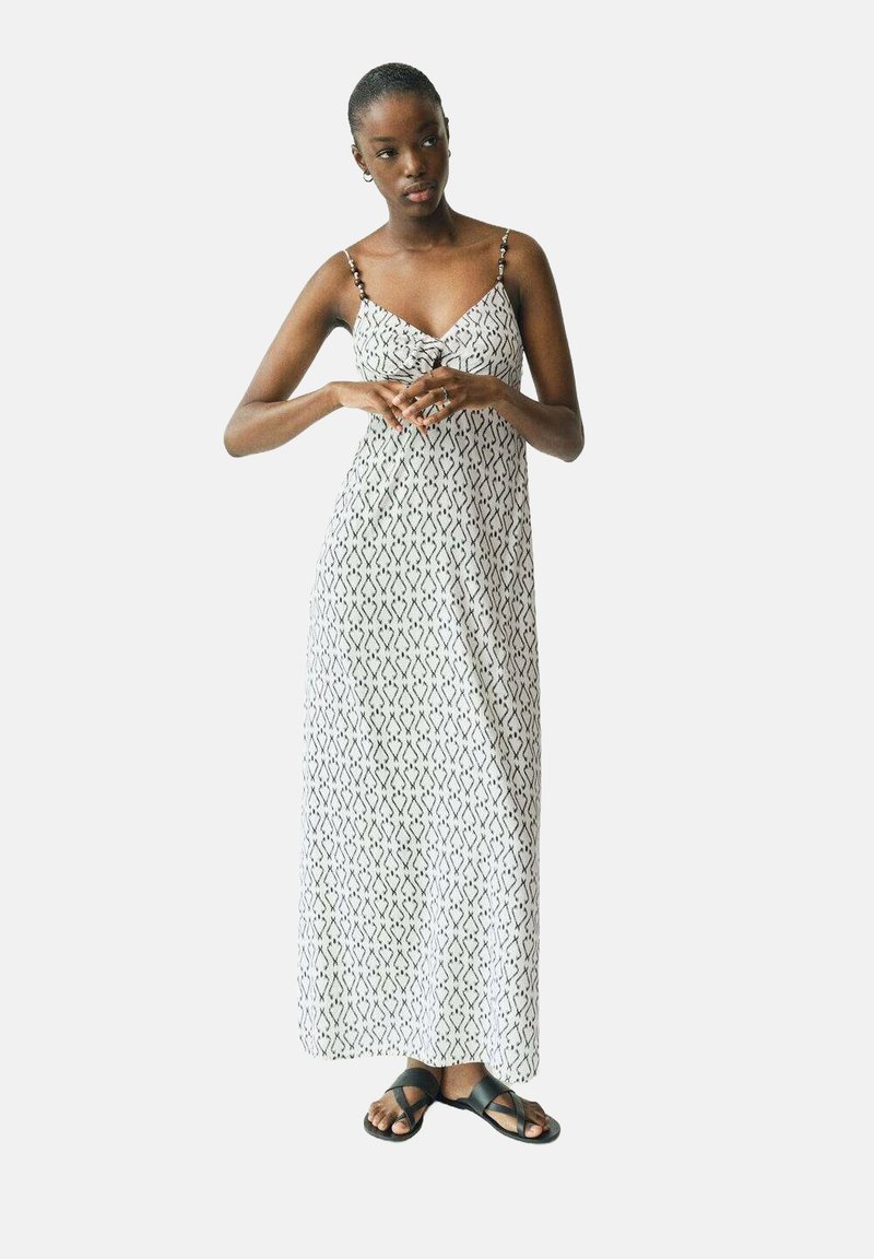 Maxikleid in Weiß mit einem schwarzen geometrischen Muster. Verstellbare Spaghetti-Träger und ein luftiges Design. Kombiniert mit schwarzen Sandalen.