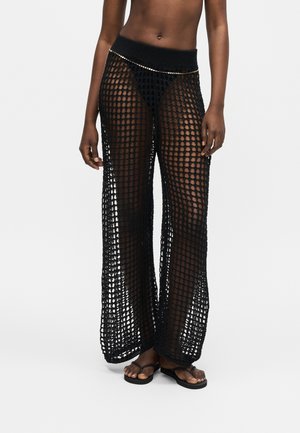 Schwarze, weit geschnittene Häkelhose, getragen über schwarzen Slips, kombiniert mit einem dünnen Kettengürtel und schwarzen Flip-Flop-Sandalen an einem stehenden Model.