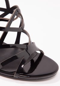 Sandale en cuir verni noir avec multiples fines brides croisées sur les orteils et la cheville, semelle visible et design à bout ouvert sur fond blanc.