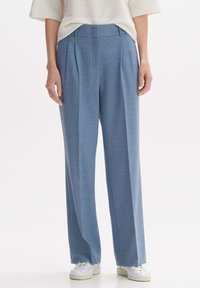 Pantalons larges bleu clair en tissu texturé avec pinces à l'avant et coupe ajustée, associés à des baskets blanches.