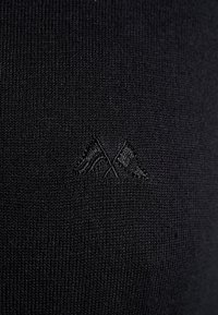 Musta neulottu kangas, jossa on hienovarainen tekstuuri, ja sen alakulmassa on kohokuvioitu malli risteävistä lipuista.