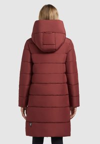 Abrigo acolchado rojo con capucha, que presenta costuras horizontales, mangas largas y un detalle triangular de marca cerca del dobladillo. Textura suave y mate.