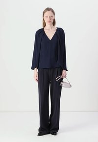 Blusa blu navy con scollatura a V, maniche lunghe e morbidi drappeggi; abbinata a pantaloni larghi blu navy e una piccola borsa grigia chiaro.