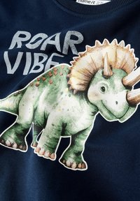 Felpa blu navy con un dinosauro verde stampato con punte marroni, testo "ROAR VIBE" in bianco sopra la grafica, tessuto morbido al tatto.