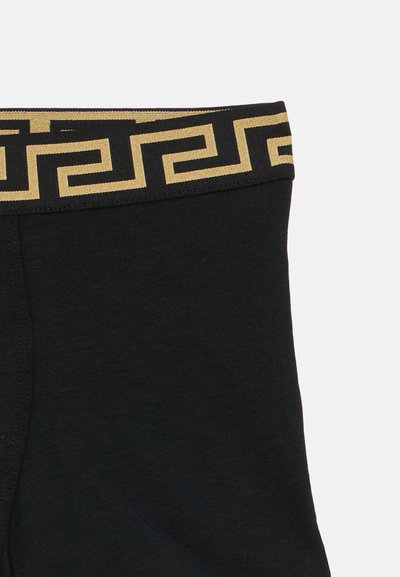 Melni kokvilnas biksītes ar zelta un melnu grieķu atslēgas rakstu vidukļa joslā. Gluda auduma tekstūra ar ciešu piegulumu un pastiprinātām šuvēm.