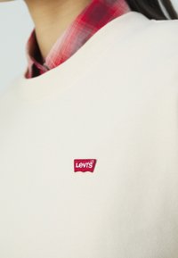 Gros plan sur une personne portant un sweat crème avec un écusson logo Levi's rouge, par-dessus le col d'une chemise à carreaux rouge.