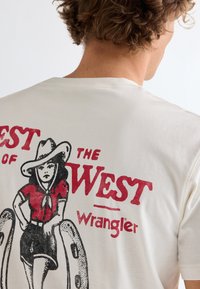 Άτομο με σγουρά μαλλιά που φοράει λευκό μπλουζάκι με κόκκινο και μαύρο σχέδιο καουμπόι "Best of the West" και λογότυπο Wrangler στην πλάτη.