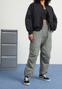 Zwarte bomberjack, grijze cargobroek met pockets en zwarte sneakers. Geweven stof, ontspannen pasvorm en monochrome kleurenschema.