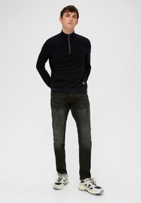 Czarny sweter z zamkiem pod szyją o ribbowanej fakturze, w zestawieniu z dopasowanymi ciemnoszarymi dżinsami i jasnymi trampkami. Prosty design z minimalnymi akcentami.