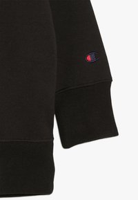 Manica di una felpa nera con polsino a costine e piccolo logo Champion ricamato in blu navy e rosso vicino al polso.