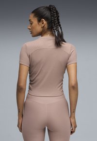 Γυναίκα που φαίνεται από πίσω να φοράει μωβ εφαρμοστό κοντομάνικο τοπ και κολάν, με τα μαλλιά σε τρία πλεξούδες δεμένα πίσω, με ορατά τατουάζ στο λαιμό και τα χέρια.