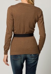 Brauner Pullover mit langen Ärmeln und rundem Halsausschnitt, weicher Textur und einem schwarzen Bundakzent. Getragen mit blauen Jeans mit Rückentaschendetails.