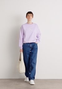 Lilla sweatshirt med fleece-tekstur og en hvit logo, kombinert med løstsittende blåjeans og hvite joggesko. Modellen holder en beige tote-bag.