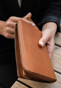 Tan leather wallet with a zipper closure, embossed logo on the front, and smooth texture. Held in a hand against a wooden surface.