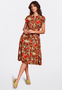 Red and beige patterned dress with short sleeves, a collar, and a tied waist. The fabric has a smooth texture, complemented by yellow sandals.