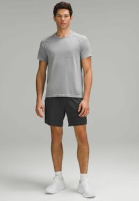 Grå texturerad kortärmad t-shirt kombinerad med svarta shorts. Modellen har på sig vita sportskor och står framför en neutral bakgrund.