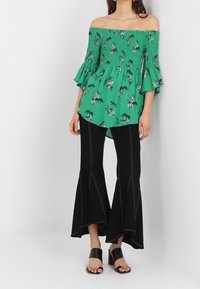 Top verde senza spalle con stampa floreale, maniche a rouches e corpetto arricciato abbinato a jeans neri a zampa con cuciture a contrasto.