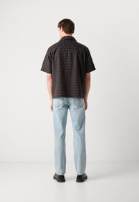 Camisa de manga corta en negro con un patrón de logo marrón repetido, combinada con jeans rectos de color azul claro y zapatos negros, vista desde atrás.