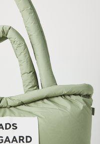 Sac fourre-tout vert rembourré avec de poignées épaisses et texturées et un haut structuré. Présente une étiquette blanche avec un texte noir attachée sur le côté.