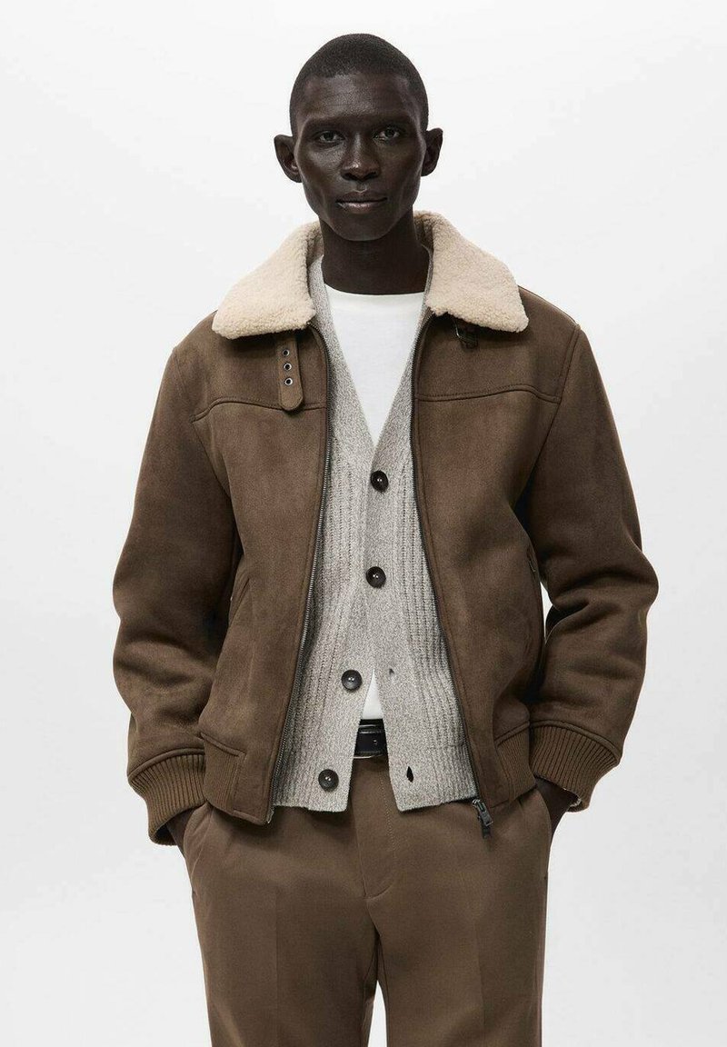 Bomber jacket in suede marrone con colletto in shearling crema, indossato sopra un cardigan grigio chiaro e una camicia bianca, abbinato a pantaloni marroni.