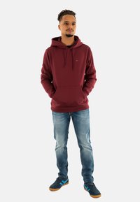 Bordeaux hoodie met een voorzak, trekkoord en een klein logo, gecombineerd met lichtblauwe jeans en donkere sneakers met blauwe accenten.