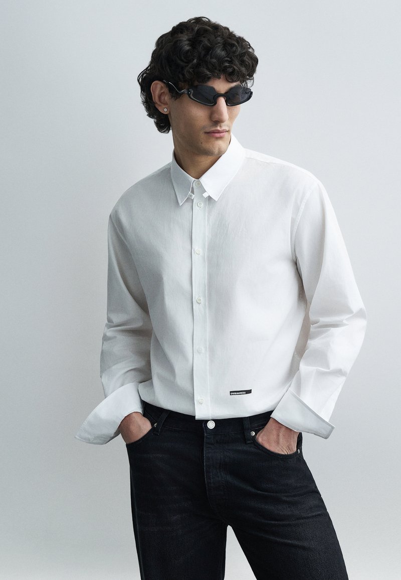 Mand med krøllet hår iført sorte solbriller, hvid skjorte med knapper og DSQUARED2-label, og sorte jeans, hænderne i lommerne, kigger til siden.