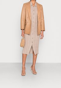 Blazer double boutonnage en cuir tan porté sur une robe tricot beige à boutons et à côtes. Les accessoires comprennent un sac à main tissé et des talons nus à lanières.