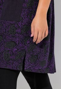 Tissu noir et violet à motifs avec des tourbillons et des triangles, présentant une fente à l'ourlet. Une main est visible, reposant sur le vêtement.