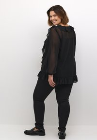 Sort bluse med flæse detaljer ved skuldre og kant, parret med sorte slim-fit jeans og blanke sorte sko. Glat, let tekstur.